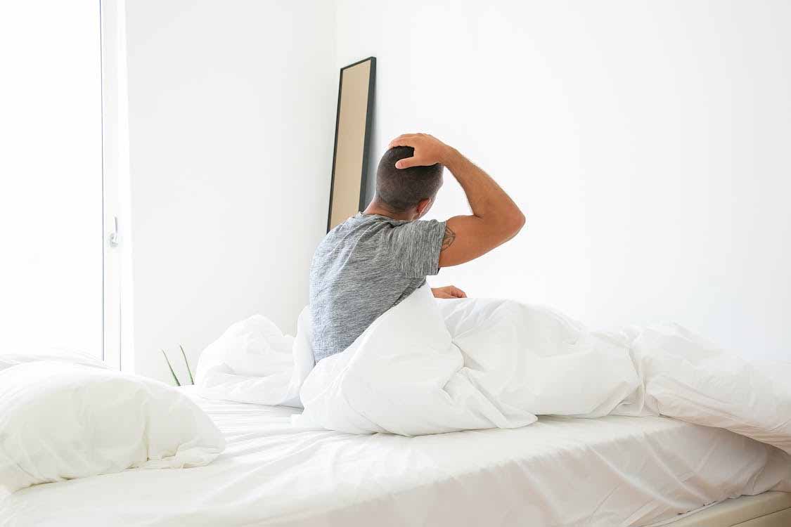 Man sitting on bed in the morning holding his head, reflecting concern about erectile dysfunction or sexual health issues